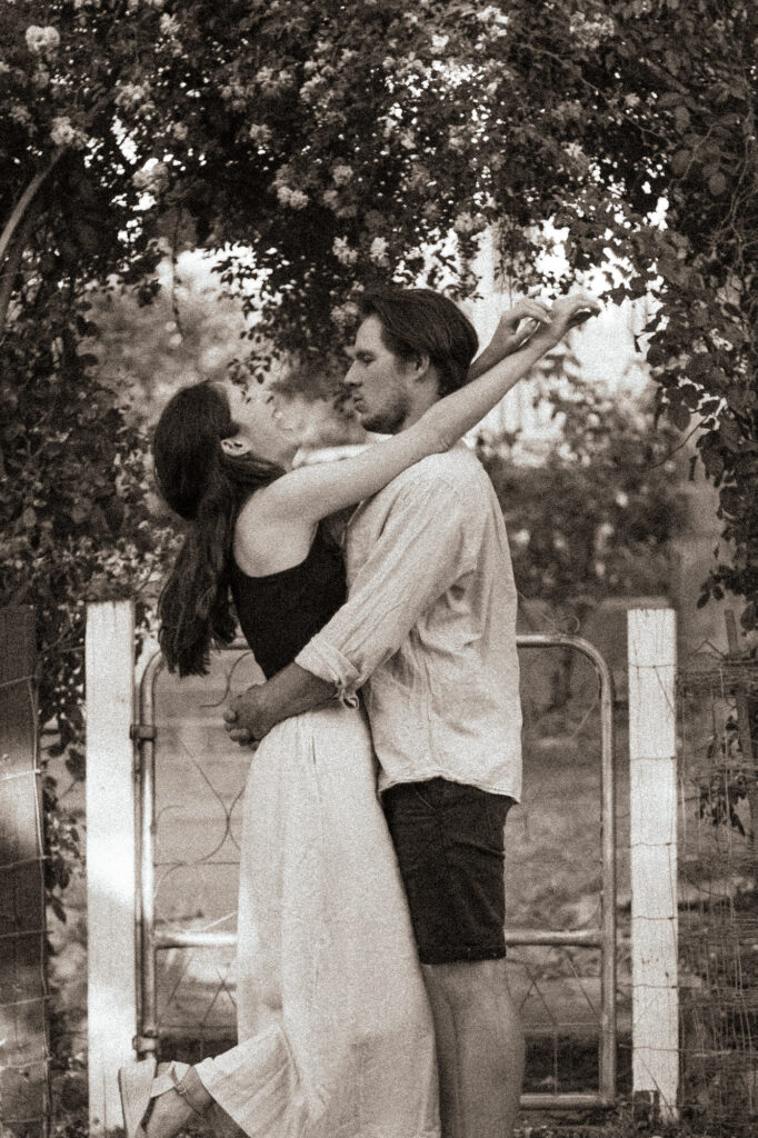 Black-and-white-of-couple-standing-in-front-of-arch-with-arms-around-each-other-her-head-thrown-back-laughing-and-her-foot-popped