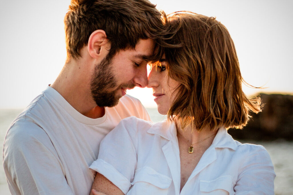 Couple-wearing-white-forehead-toforehead-and-nose-to-nose-at-the-beach-with-the-sunlight-behind-them