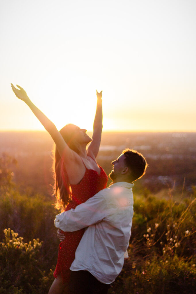 Couple-at-sunset-man-lifting-woman-up-as-throws-her-hands-towards-the-sky-soaking-in-the-warmth-and-enjoying-the-moment