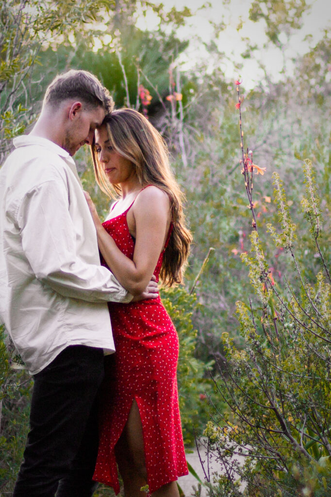 Couple-standing-close-together-on-small-bush-path-with-foreheads-pressed-together-surrounded-by-greenery-and-native-flowers