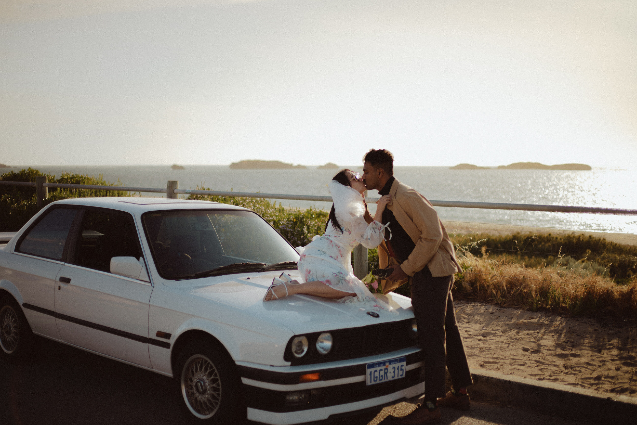 Couple sitting on car bonnet kissing with beach in background